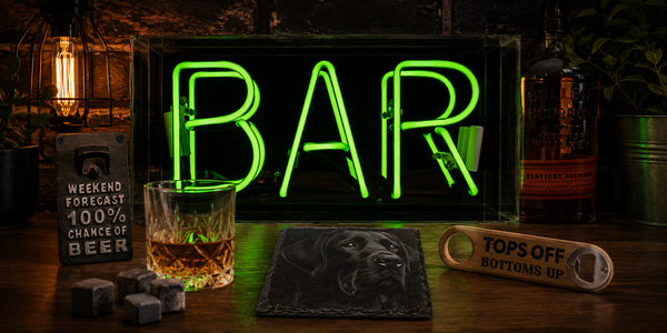 Home and bar collection banner featuring a green BAR neon sign, whisky glass, slate dog coaster, bottle opener and bar accessories in a warm moody interior setting