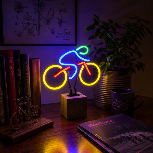 Colourful bicycle neon lamp on a desk beside books and plants with neon light reflections in a moody interior