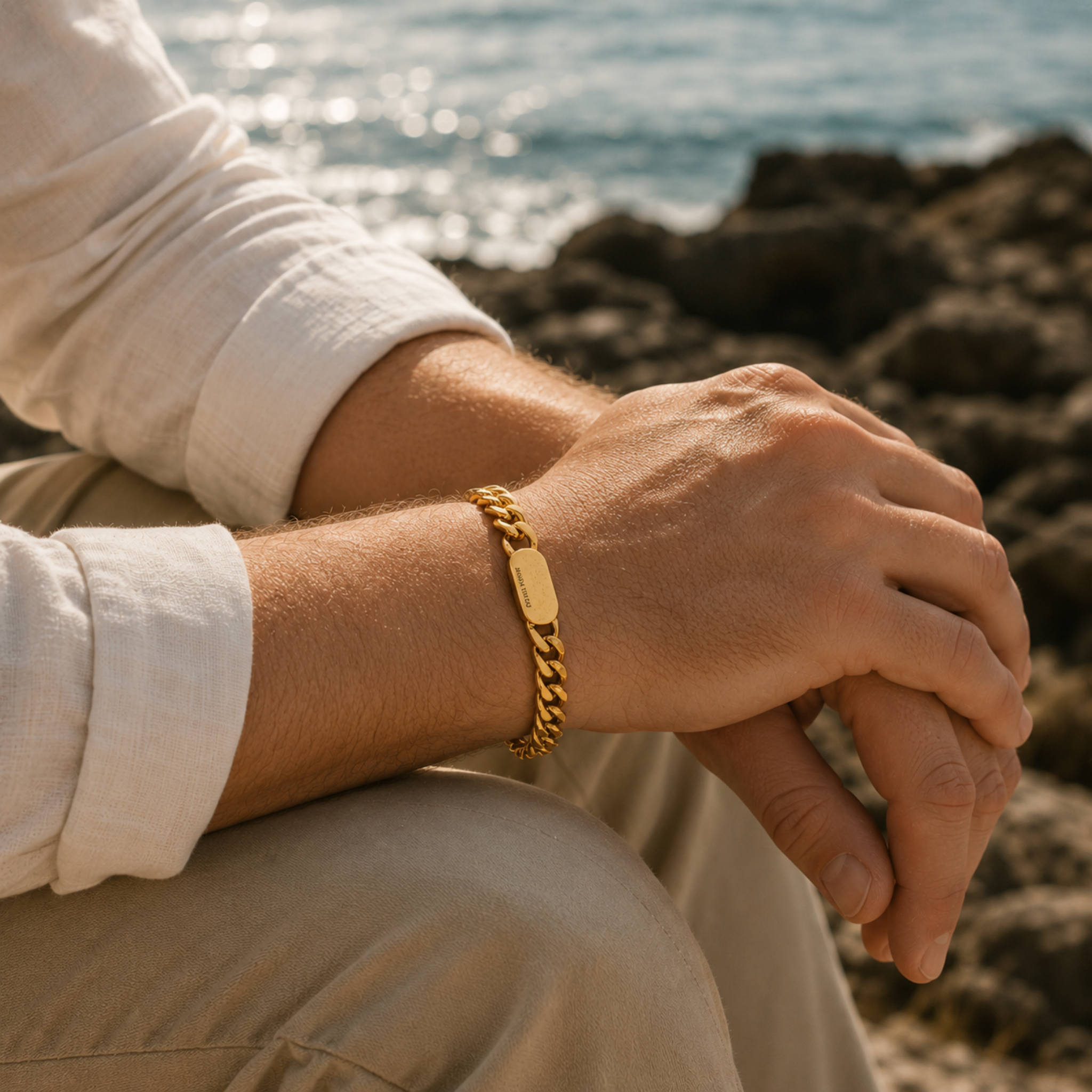 Man wearing a gold colour stainless steel curb chain bracelet while relaxing on coastal rocks beside the sea in warm natural sunlight.