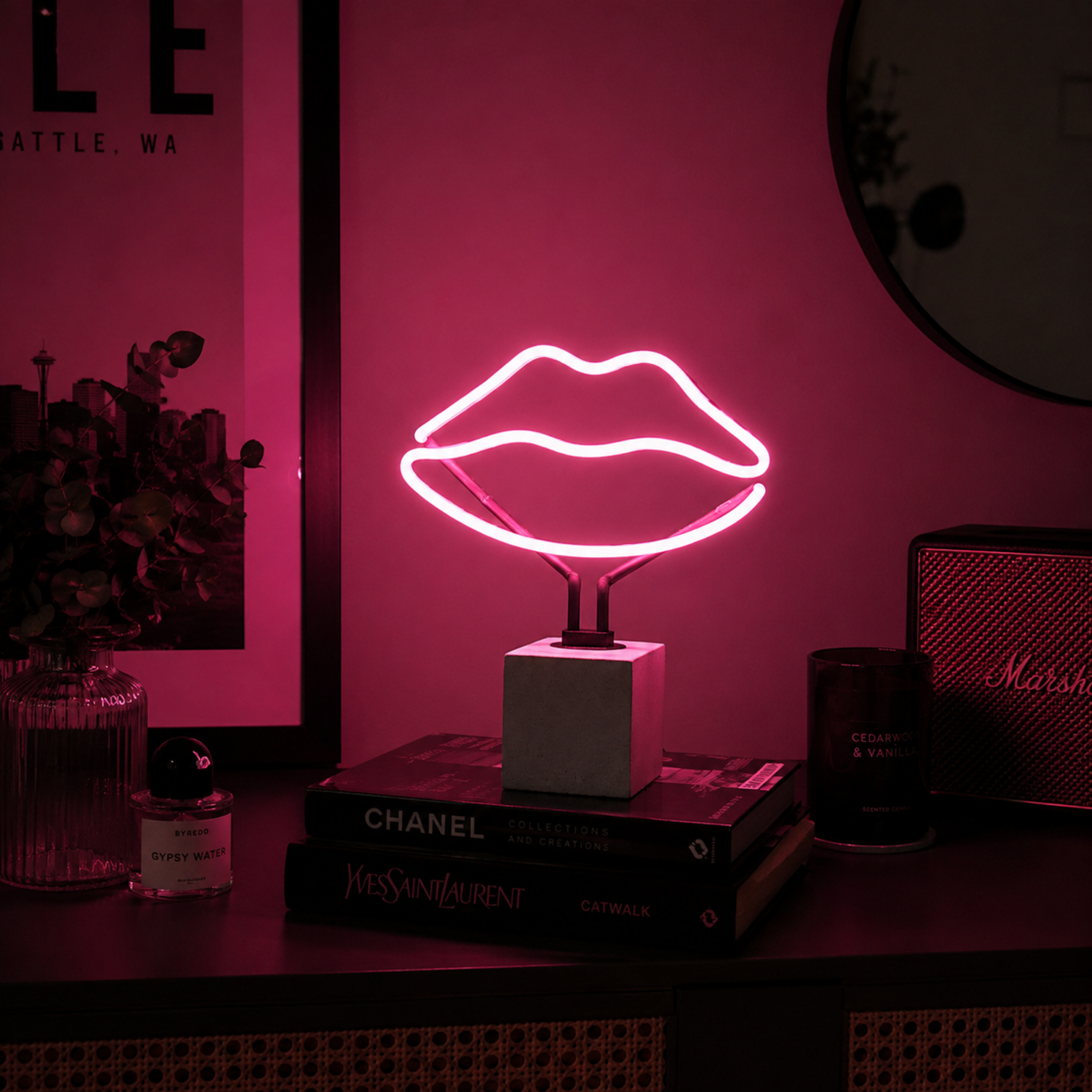 Pink lips neon lamp glowing beside books and speaker in a moody interior setting