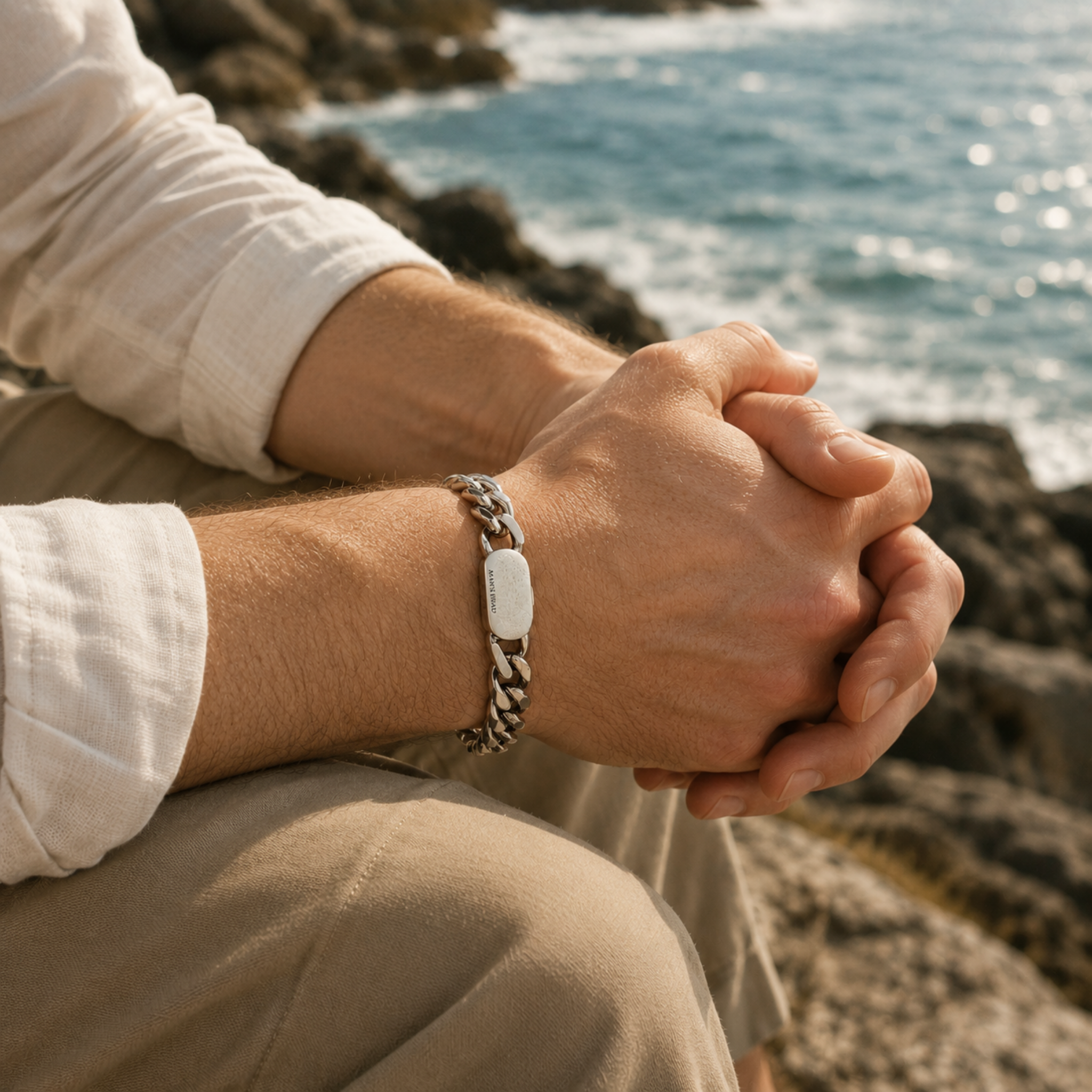 Man wearing a stainless steel curb chain bracelet while sitting on coastal rocks overlooking the sea in warm natural sunlight.