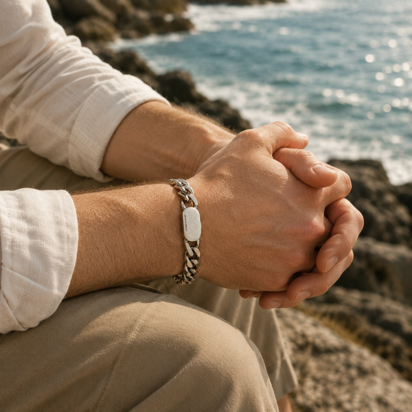 Man wearing a stainless steel curb chain bracelet while sitting on coastal rocks overlooking the sea in warm natural sunlight.