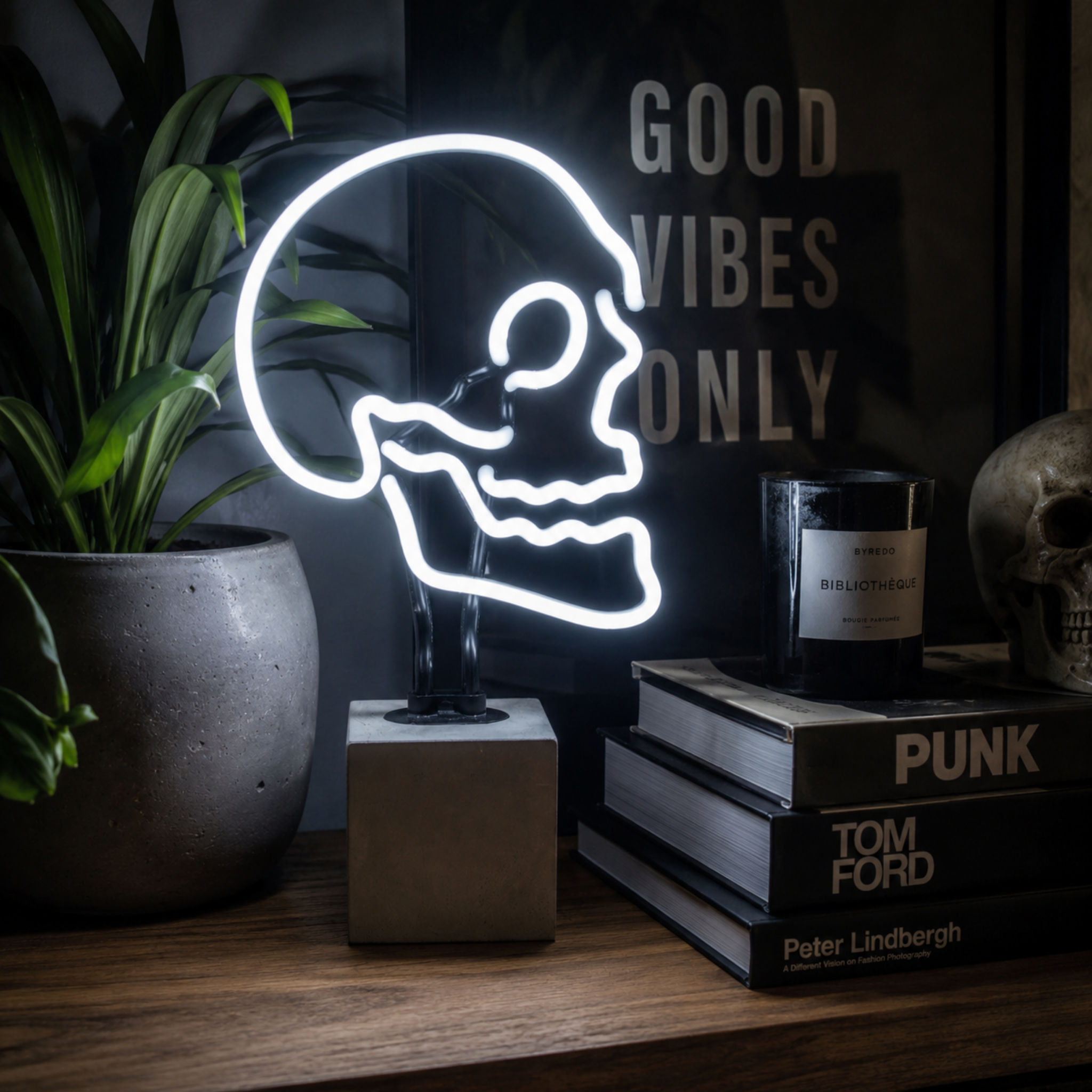 White skull neon lamp glowing on a wooden shelf beside books, candle and indoor plant in a moody interior setting